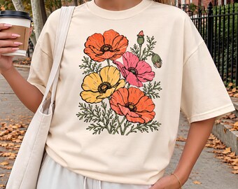 May include: An ivory short-sleeved t-shirt featuring a central graphic print of four blooming poppies in shades of orange, yellow, and pink, accompanied by green foliage and buds. The shirt has a crew neckline and a relaxed, loose-fitting silhouette.