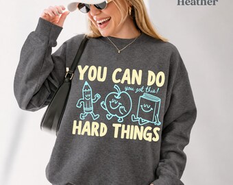 May include: A dark heather gray crewneck sweatshirt featuring the text YOU CAN DO HARD THINGS and you got this! with light blue line art illustrations of a smiling pencil, an apple, and a book. The design is printed in yellow and light blue.