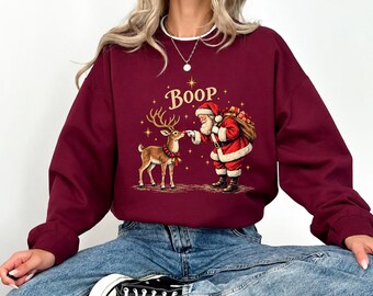 May include: A maroon long-sleeve sweatshirt featuring a graphic of Santa Claus touching the nose of a reindeer. The text "Boop" is printed above the figures in gold lettering, surrounded by small gold stars. The sweatshirt has a white crew neckline.
