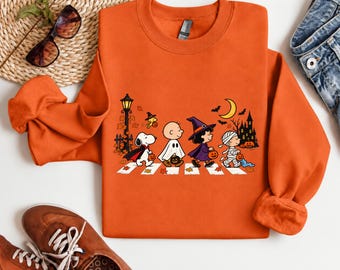 May include: An orange long-sleeve sweatshirt featuring a graphic print of Peanuts characters dressed in Halloween costumes, including Snoopy as a vampire, Charlie Brown as a ghost, Lucy as a witch, and Linus as a mummy, walking on a crosswalk.