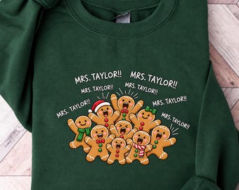 May include: A dark green crewneck sweatshirt featuring a graphic of nine gingerbread cookies with various accessories like scarves and hats. The text "MRS. TAYLOR!!" is printed multiple times in white above the gingerbread characters.