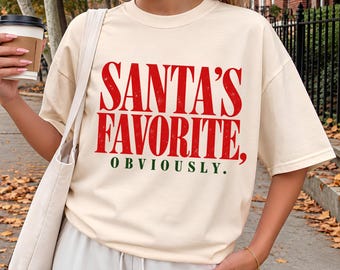May include: An ivory short-sleeve t-shirt featuring the red text SANTA'S FAVORITE, and the green text OBVIOUSLY. The shirt has a crew neckline and a relaxed fit, with a distressed texture on the red lettering.