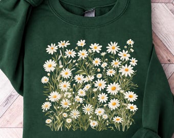May include: A dark forest green crewneck sweatshirt featuring a central graphic print of a dense field of white daisies with yellow centers and green stems and leaves. The long-sleeved garment has a ribbed collar, cuffs, and hem.