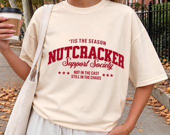 May include: An ivory short-sleeve t-shirt featuring red text that reads 'TIS THE SEASON NUTCRACKER Support Society NOT IN THE CAST STILL IN THE CHAOS with small red stars on either side of the bottom line of text.
