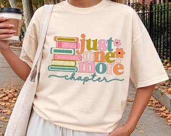 May include: An ivory t-shirt featuring a graphic of a stack of five colorful books and the text "just one more chapter" in multi-colored lettering, accompanied by two small flower illustrations in pink and orange.