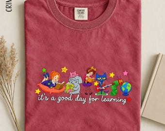 May include: A crimson short-sleeve t-shirt featuring a graphic of various storybook characters reading and playing music, with the text "it's a good day for learning" printed below. The shirt has a Comfort Colors brand label at the neck.