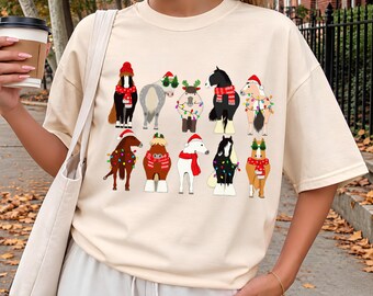 May include: An ivory short-sleeved t-shirt featuring a graphic print of ten horses decorated for the holidays. The horses are wearing Santa hats, red scarves, and are wrapped in colorful string lights against a plain light background.