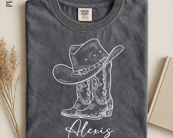 May include: A charcoal gray crew neck t-shirt featuring a white line art graphic of a cowboy hat resting on a pair of western boots. The name Alexis is printed in white script below the boots. The shirt has a Comfort Colors brand label.