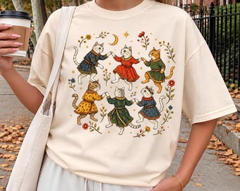 May include: An ivory-colored t-shirt featuring a graphic print of six cats wearing colorful tunics and holding hands in a circle. The design includes small floral elements, a crescent moon, and stars surrounding the dancing cats.