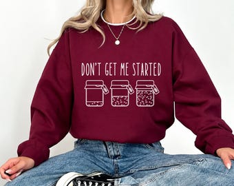 May include: A maroon long-sleeve sweatshirt featuring a white graphic of three jars with varying levels of contents and the text "DON'T GET ME STARTED" printed above them. The sweatshirt has a crew neckline with a white trim detail.