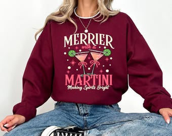 May include: A maroon crewneck sweatshirt featuring a graphic of two martini glasses with lime garnishes. The text reads "MERRIER with a MARTINI Making Spirits Bright" in cream and pink lettering, accented by small pink stars and white dots.