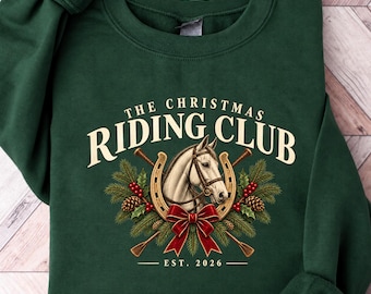 May include: A dark green crewneck sweatshirt featuring a graphic of a horse head inside a horseshoe, surrounded by pine branches, pinecones, and red berries. The text reads THE CHRISTMAS RIDING CLUB EST. 2026 in cream-colored lettering.