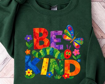 May include: A dark green long-sleeve sweatshirt featuring a colorful graphic print. The design includes the text BE KIND in large, textured, multi-colored letters, a green caterpillar, a multi-colored butterfly, and various small flowers.
