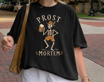May include: A black short-sleeve t-shirt featuring a graphic of a skeleton wearing a hat and shorts, holding a beer mug. The text PROST is printed above the skeleton and MORTEM is printed below, with small leaf accents on either side of the word.