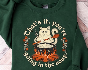 May include: A dark green crewneck sweatshirt featuring a graphic of a cat stirring a black cauldron over a campfire. The text reads, "That's it, you're going in the soup." The design includes orange flowers and flames, with a cream-colored cat holding a wooden spoon.