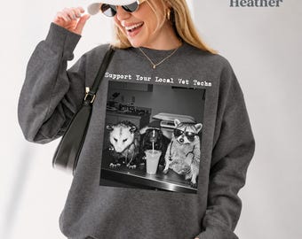 May include: A dark heather gray crewneck sweatshirt featuring a black and white graphic print of an opossum, a skunk, and a raccoon wearing stethoscopes and sunglasses. The text above the graphic reads Support Your Local Vet Techs.