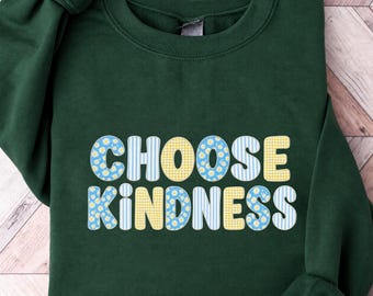 May include: A forest green crewneck sweatshirt featuring the text CHOOSE KINDNESS in large, rounded letters. The letters are filled with alternating patterns of yellow gingham, blue and white stripes, and small white daisies on blue.