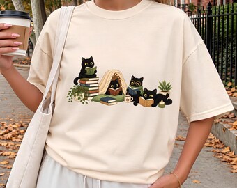 May include: An ivory t-shirt featuring a graphic print of four black cats reading books. One cat sits on a stack of books, one is inside a small tent, one wears glasses while reading, and one lies down with a book. A potted plant and candle are included.