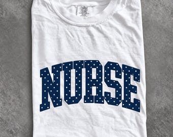 May include: A white short-sleeved t-shirt featuring the word NURSE printed in a dark navy blue collegiate font with a white polka dot pattern. The shirt has a crew neckline and a Comfort Colors brand label visible at the inner collar.