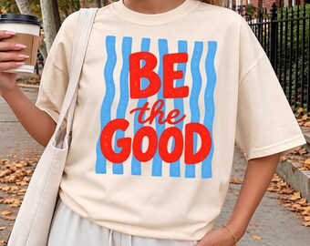 May include: An ivory short-sleeved t-shirt featuring a graphic print with vertical blue wavy lines and the red text "BE the GOOD" centered on the front. The shirt has a crew neckline and a relaxed fit, paired with a matching ivory tote bag.