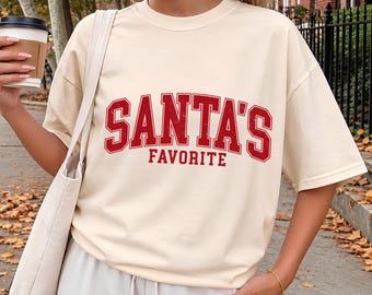 May include: An ivory short-sleeved t-shirt featuring the red text SANTA'S FAVORITE in a distressed collegiate font across the chest. The shirt has a crew neckline and a relaxed fit, paired with a matching ivory tote bag.