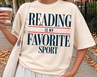 May include: An ivory short-sleeved t-shirt featuring the dark blue text "READING IS MY FAVORITE SPORT" with two horizontal red lines separating the words. The shirt has a crew neckline and a relaxed fit, made of a solid-colored fabric.