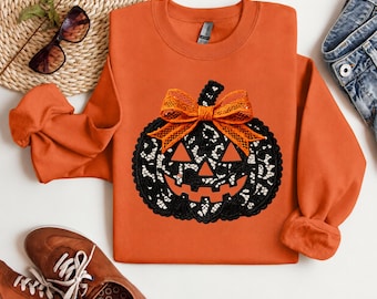 May include: An orange long-sleeve sweatshirt featuring a black jack-o'-lantern graphic with a white leopard print pattern inside the pumpkin shape. The pumpkin is decorated with a bright orange mesh bow at the top center.