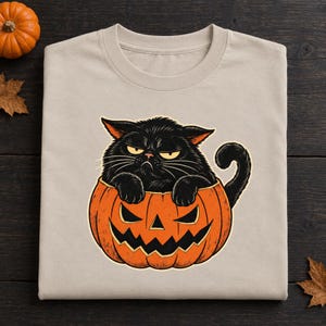 May include: A beige t-shirt featuring a graphic illustration of a grumpy black cat sitting inside an orange jack-o'-lantern pumpkin. The cat has yellow eyes and long whiskers, while the pumpkin displays a carved, jagged, and toothy grin.