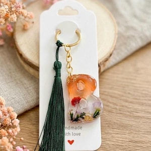 May include: A keychain featuring a resin letter B with an orange-to-clear gradient, embedded with dried flowers and gold flakes. It includes a dark green tassel and a gold-tone metal ring attached to a white display card with the text handmade with love.