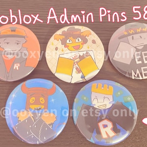 May include: Five circular pin-back buttons featuring Roblox character illustrations. The pins measure 58mm in diameter. The designs include characters with orange, yellow, grey, blue, and purple colour schemes, some with text reading MEET ME.