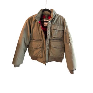 May include: A sage green bomber jacket featuring a high collar, a front zipper closure, and multiple utility pockets on the chest and sleeves. The interior is lined with a red and black buffalo plaid fabric, and the cuffs and hem are ribbed.