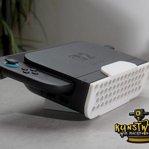 Puede incluir: Una consola Nintendo Switch negra colocada en un soporte blanco impreso en 3D con un panel frontal de patrón de panal. La consola tiene un joystick azul en el mando izquierdo y el logotipo de Nintendo Switch es visible en el centro.