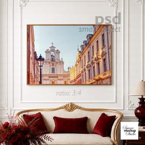 May include: A rectangular wall art print featuring a European street scene with a church and historic buildings, displayed in a gold frame on a white panelled wall above a cream-coloured sofa with three red velvet throw pillows and a lamp.