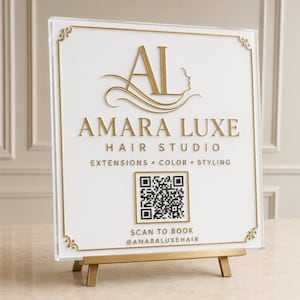 May include: A square white acrylic sign with gold lettering and a gold border on a gold easel. The sign features the text AMARA LUXE HAIR STUDIO, EXTENSIONS, COLOR, STYLING, SCAN TO BOOK, @AMARALUXEHAIR, a logo with the letters AL, and a QR code.
