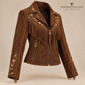 Puede incluir: Una chaqueta marrón con largos flecos en el pecho y las mangas, bordados florales, detalles de tachuelas metálicas y bolsillos con cremallera. La chaqueta incluye un cierre frontal de cremallera y el texto RODEO DRIVE LEATHER HANDCRAFTED WESTERN GOODS.
