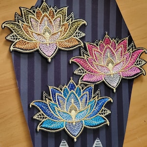 May include: Three embroidered lotus flower patches featuring intricate stitching in metallic gold, pink, blue, and bronze threads. Each patch has a pointed petal design with a black border and detailed filigree accents at the base of the petals.