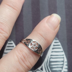 May include: A silver-toned metal ring featuring a central pentagram design flanked by intricate openwork knot patterns. The band has a polished finish and a continuous cutout style that wraps around the finger, showcasing a detailed symmetrical motif.