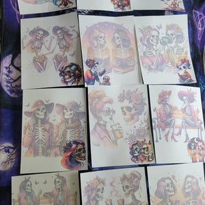 May include: A collection of rectangular sheets featuring colourful illustrations of skeletons with vibrant hair, wearing hats, glasses, and patterned clothing. The designs depict skeletons in various social settings, including sitting on benches, drinking beverages, and playing music.