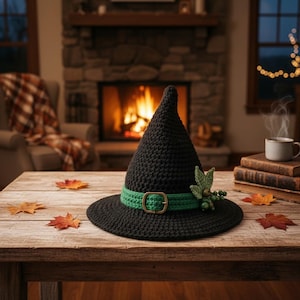 May include: A black crocheted witch hat featuring a green band with a metallic buckle. The hat is adorned with a small cluster of green crocheted leaves and berries attached to the side of the band, resting on a wooden surface.