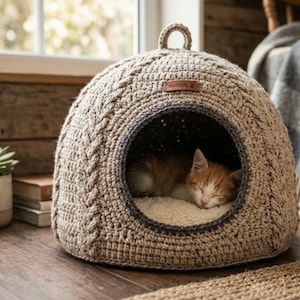 Peut inclure: Un dôme de couchage pour animal en crochet beige avec un motif torsadé et une petite boucle sur le dessus. Une étiquette en cuir marron marquée "HANDMADE" avec une icône de patte est fixée à l'avant. Un petit chaton roux et blanc dort à l'intérieur.