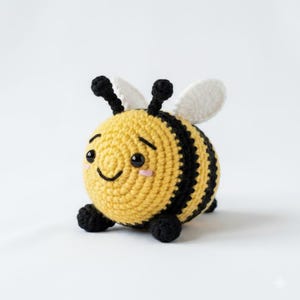 May include: A round, crocheted bee plush toy featuring a yellow and black striped body, two small black antennae, and two white wings. The bee has black safety eyes, a stitched smiling mouth, and small pink blush marks on its cheeks.