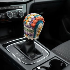 Op de afbeelding: Een gehaakte hoes voor de versnellingspook met een kleurrijk granny square patroon in oranje, teal, geel, paars en crème. Dit handgemaakte textielaccessoire is geplaatst over de zwarte lederen versnellingspook in een auto-interieur.