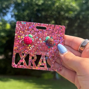 May include: A pair of small, round, textured stud earrings attached to a square, pink glitter display card. One earring is bright pink and the other is dark brown. The display card features the word XRAY cut out in large, bold, block letters.