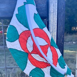May include: A crescent-shaped quilted fanny pack featuring a white base fabric with a vibrant red floral pattern and green leaf appliques. The bag includes a white zipper closure running diagonally across the front, with visible stitching.