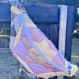 May include: A crescent-shaped crossbody bag constructed from a patchwork quilt featuring various fabric patterns in yellow, lavender, and floral prints. The bag includes a white zipper closure running diagonally across the front and a patterned strap.