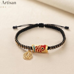 Può includere: Un bracciale in corda intrecciata nera con un ciondolo a forma di pesce koi dorato con dettagli in smalto rosso e blu, accompagnato da un ciondolo circolare dorato inciso con il carattere cinese per la buona fortuna, logo Infinite Artisan Store.