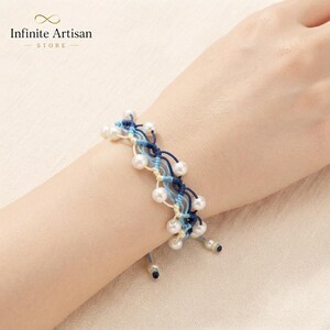 May include: A multi-strand bracelet featuring woven navy blue, light blue, and cream-colored cord. The design incorporates small, round, white pearls spaced evenly throughout the intricate knotted pattern, creating a layered and textured appearance.