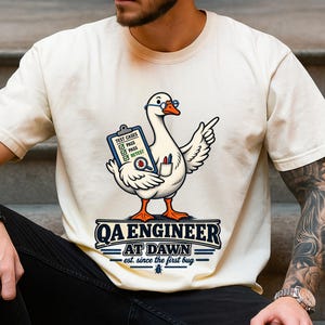May include: A cream-coloured t-shirt featuring a graphic of a white goose wearing blue glasses, holding a clipboard with the text "TEST CASES" and "PASS" checkboxes, and pointing. Below the goose, the text reads "QA ENGINEER AT DAWN est. since the first bug".