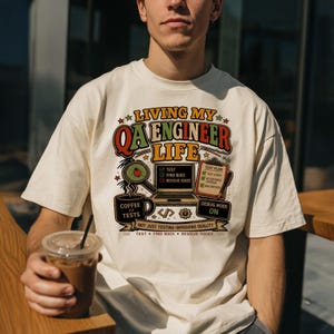 Puede incluir: Una camiseta color hueso con un gráfico que incluye el texto "LIVING MY QA ENGINEER LIFE". El diseño muestra una computadora portátil, una lupa sobre un insecto, una taza de café "COFFEE // TESTS", un portapapeles y "DEBUG MODE ON".