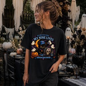 Puede incluir: Una camiseta negra con un gráfico de un gato negro con gorra azul, trabajando en un portátil con una pegatina de calavera. El diseño incluye fantasmas, una calabaza, una luna creciente y el texto "DEBUGGING MY NINE LIVES" y "DEBUGGING SINCE FOREVER."